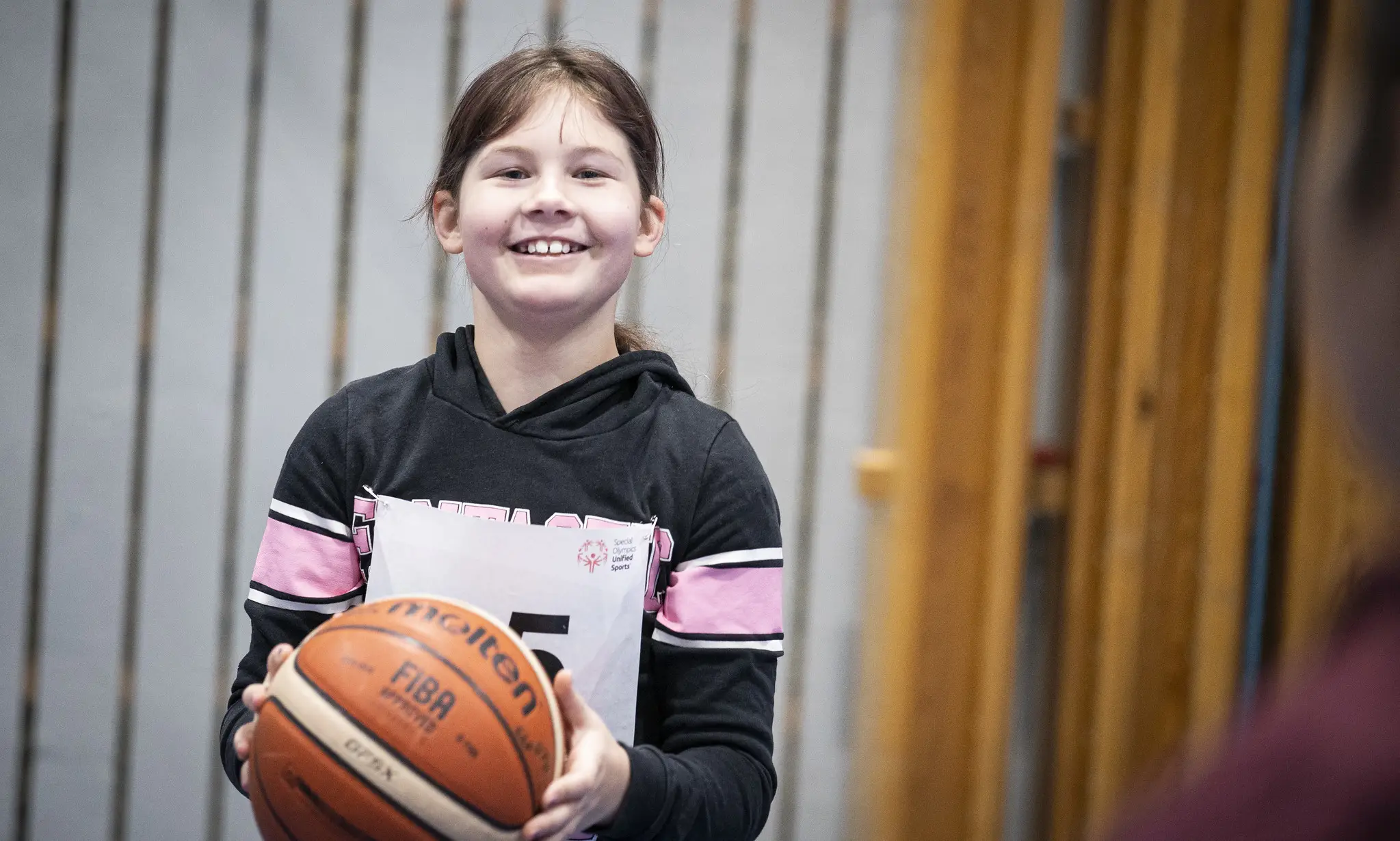 Flicka i sporthall håller i en basketboll. Hon ser glad ut och tittar mot kameran.
