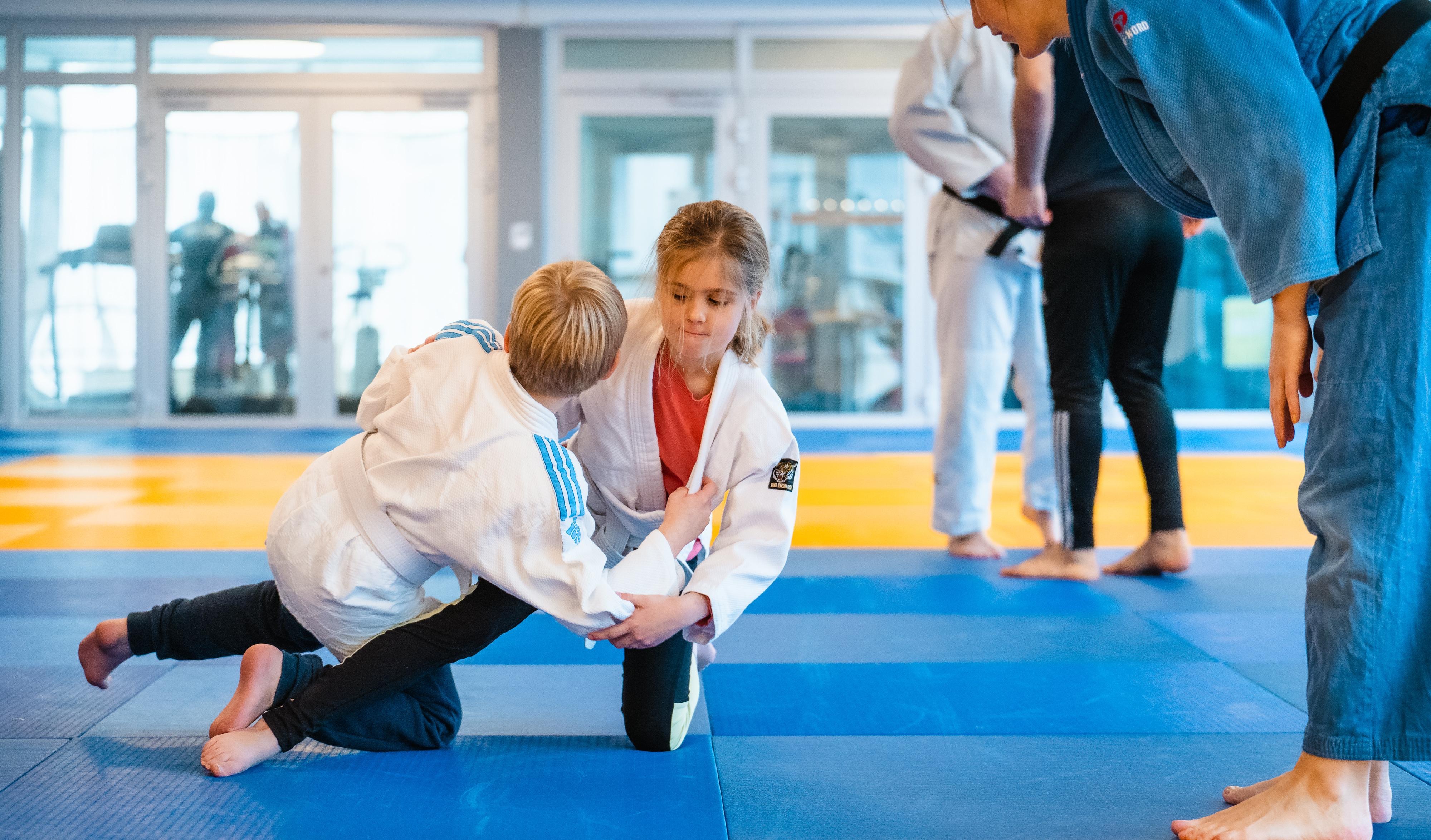 Inomhusmiljö. Två barn på en judomatta, de håller om varandra i ett grepp. Båda har vita judodräkter. Intill barnen står en ledare och ger instruktioner och peppar. 