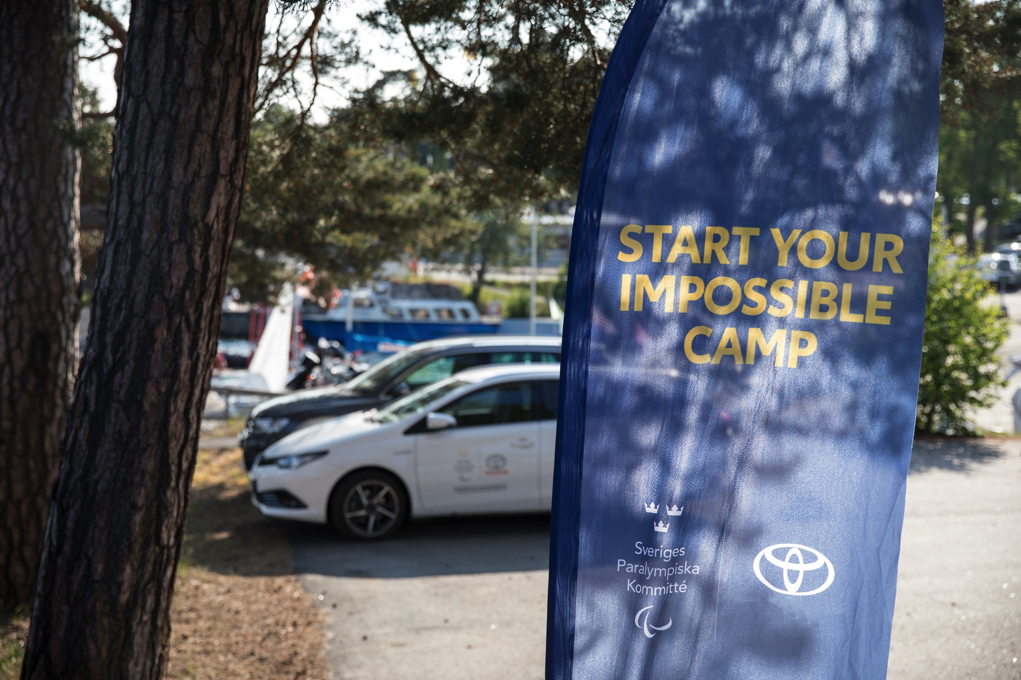 Utomhusmiljö. En parkering med Toyotabilar, i förgrunden en mörkblå flagga med texten Start Your Impossible Camp. En tall står på sidan.