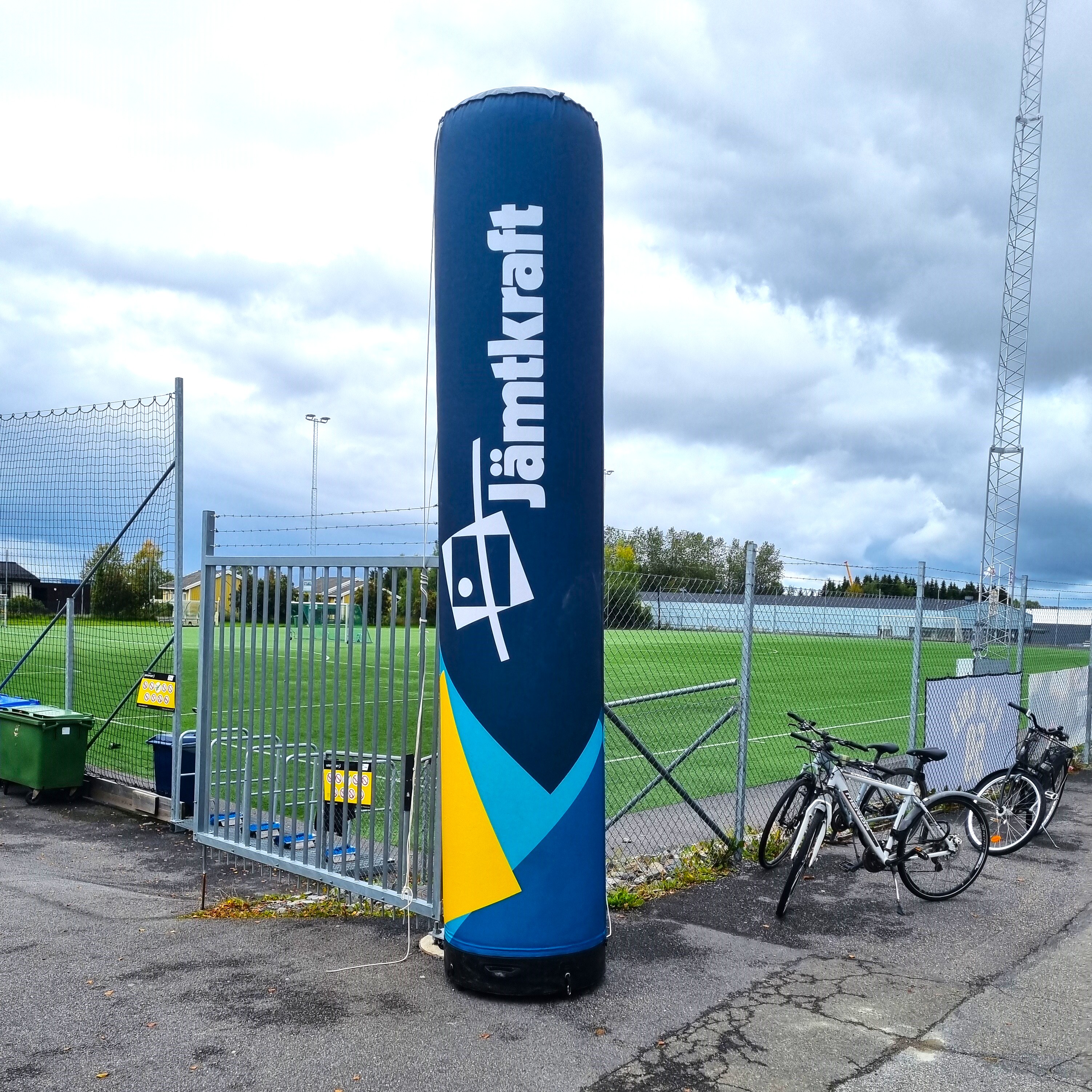 Parasportdagen. Vy över grön fotbollsplan vid Hofvallen, himlen är täckt med moln både ljusa och väldigt regntunga. Mitt i bild syns en uppblåst pelare med loggan Jämtkraft. Till höger om den, några parkerade cyklar.