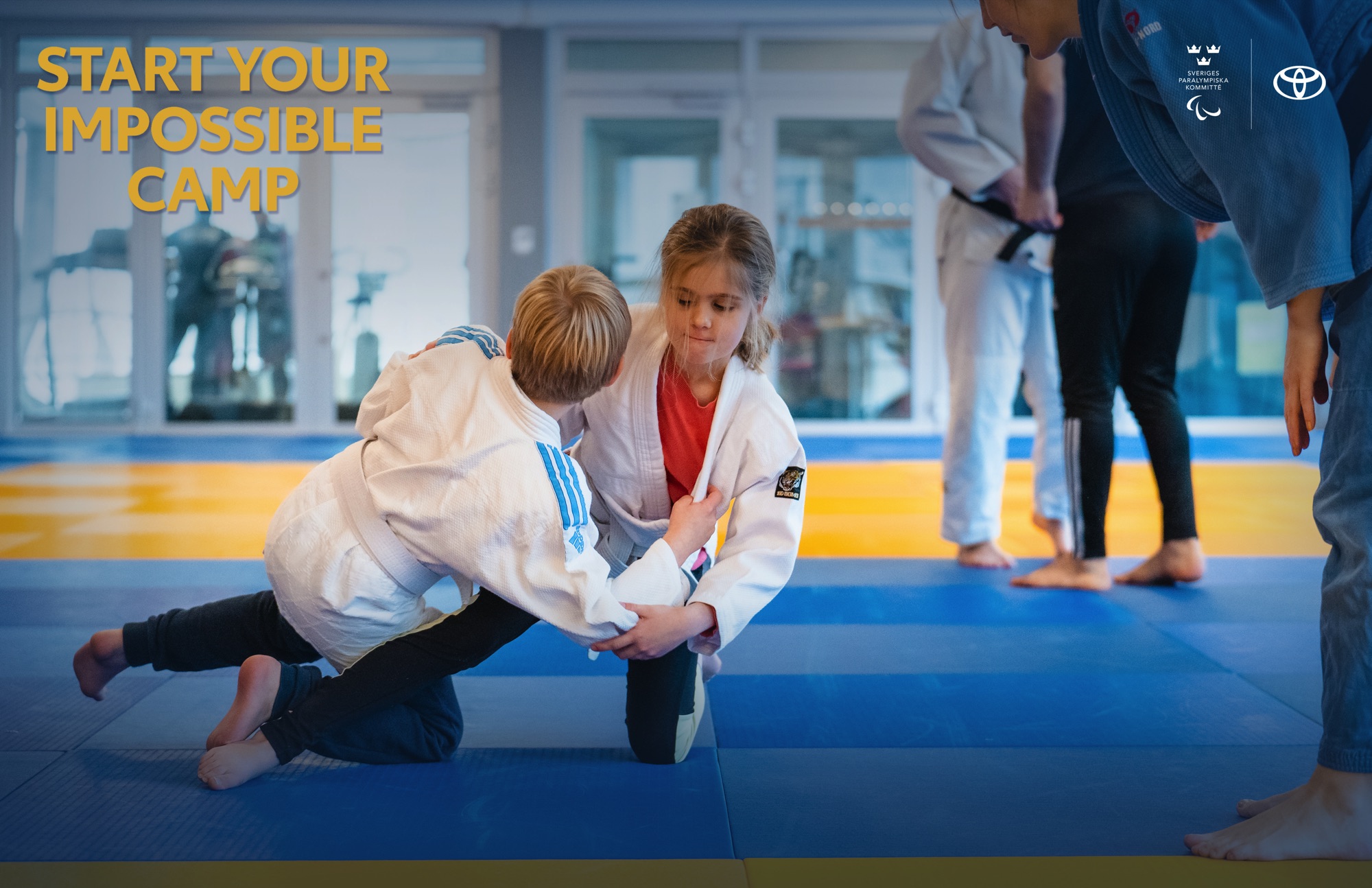 Inomhusmiljö, judosal. Två barn testar judogrepp på en judomatta. En ledare står bredvid och ger instruktioner.