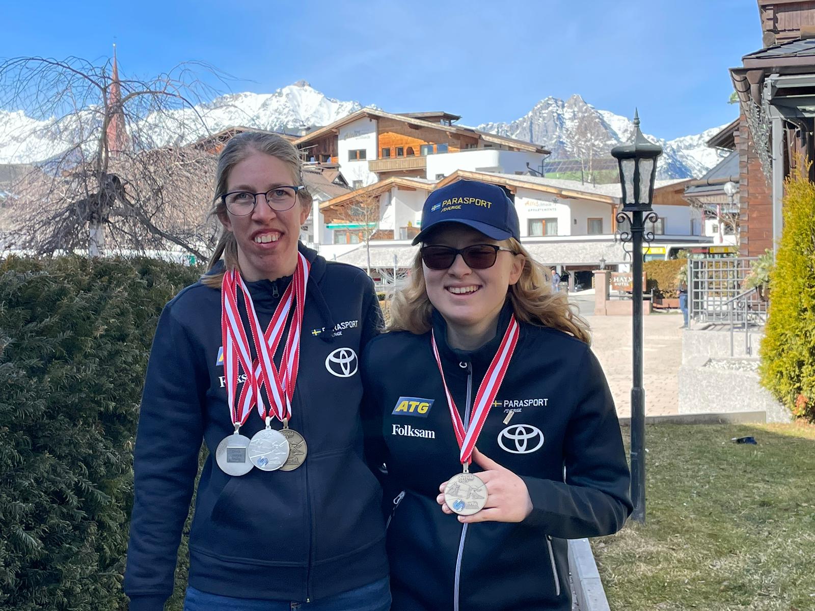 Kristin Björfeldt, längd, och Elmie Gerhardsson Danielsson, alpint, visar upp sina medaljer från skid-VM i Österrike. Bakom Elmie och Kristin syns buskar, ett hus och alptopper.