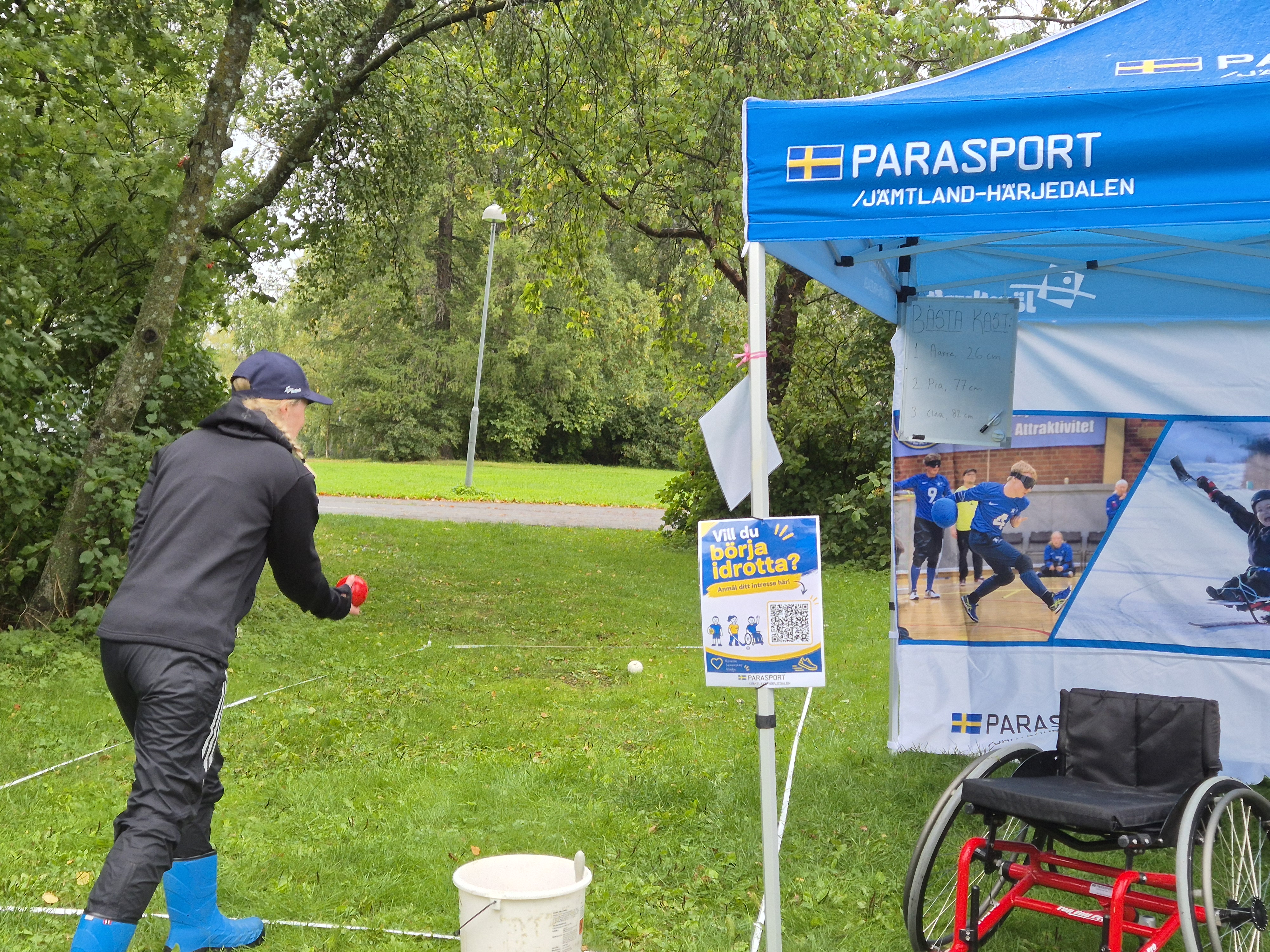 Östersund Summit 2026. Grönt gräs med ett blått Parasport-tält. Vid tältet en röd sportrullstol. Några beach-flags och inspirerande roll-ups. Till vänster om tältet står en ung kvinna och spelar boccia.