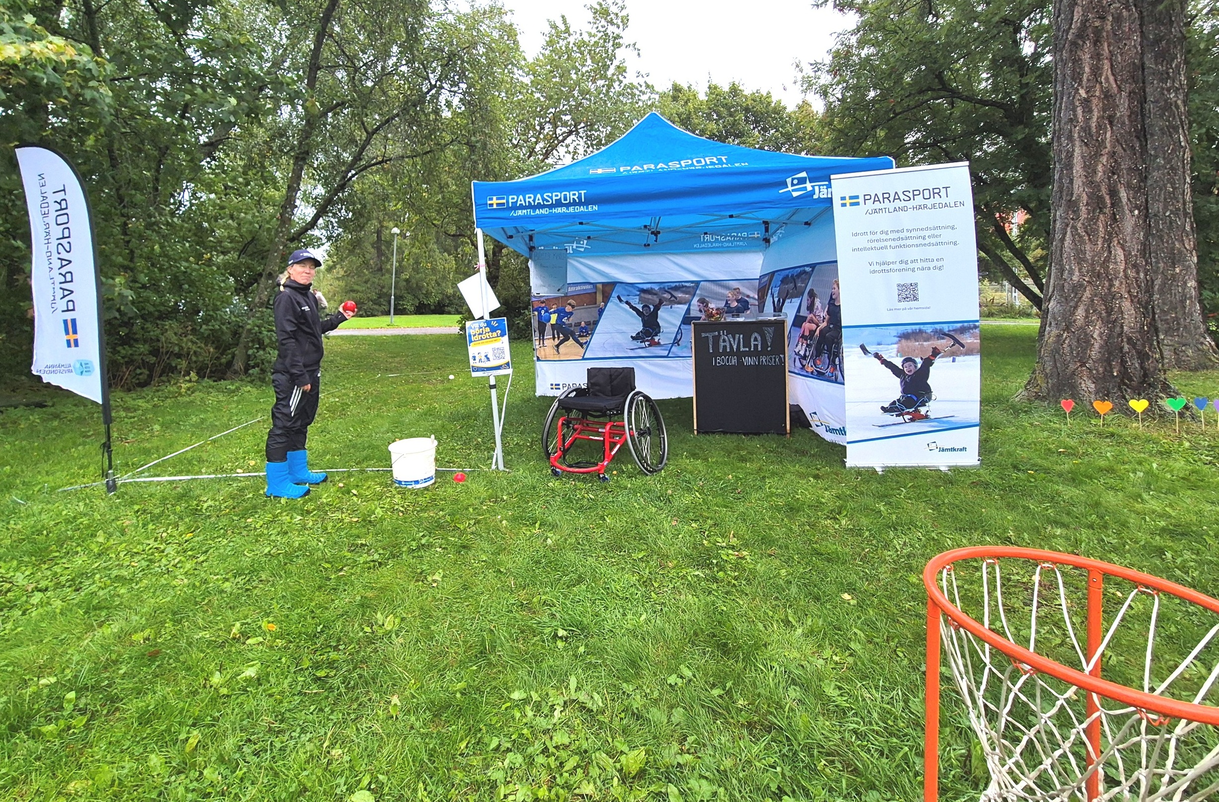Östersund Summit 2026_ I nedre högra hörnet skymtas en basketkorg. Grönt gräs med ett blått Parasport-tält. Några beach-flags och inspirerande roll-ups. Till vänster om tältet står en ung kvinna med bocciaboll i handen och tittar mot kameran.